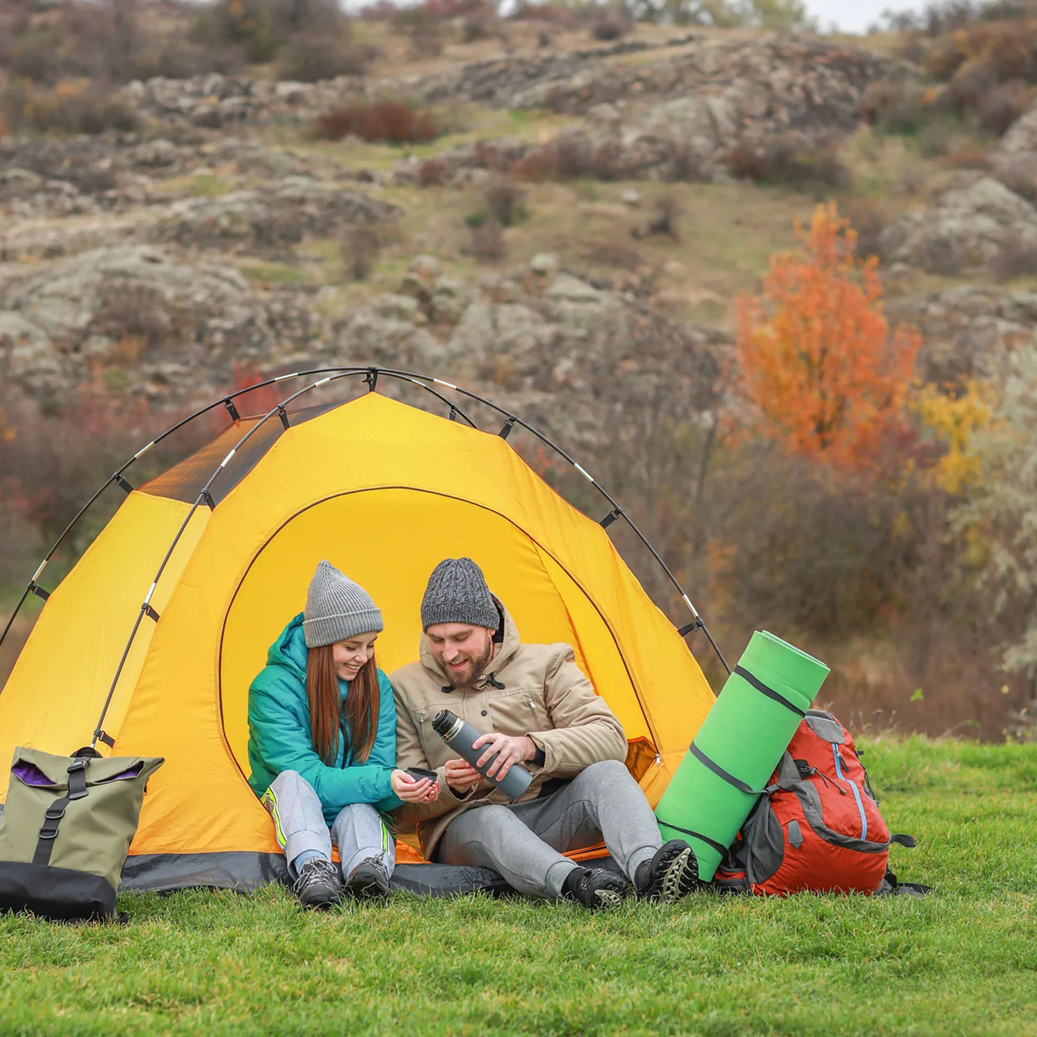Two campers in knit hats sit in a yellow tent on an autumn hillside