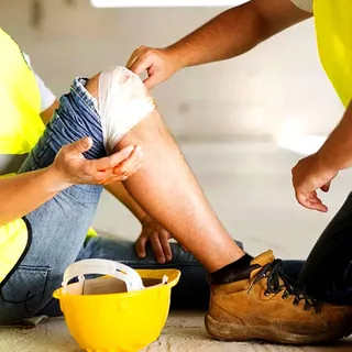 A coworker wraps a scraped knee, a yellow hard hat on the floor